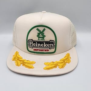 May include: Cream-colored trucker hat with a mesh back and a curved brim. The front features a green and white Heineken logo with a windmill design and the word "IMPORTED". Two embroidered yellow leaf designs adorn the brim.