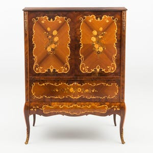 Puede incluir: Un mueble de madera con acabado marrón oscuro, con diseños florales incrustados y adornos decorativos. El mueble tiene dos puertas con marquetería floral y un cajón con detalles similares. Las patas son curvas y el estilo general es ornamentado.