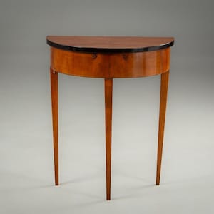 May include: A semi-circular wooden table with three slender legs. The table has a dark brown trim around the top and a light brown finish. The legs are tapered and the table is set against a grey background.