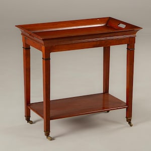 May include: A mahogany serving table with a rectangular tray top and a lower shelf. The table has four legs with small brass casters. The legs are connected by a lower shelf. The table is approximately 30 inches tall.