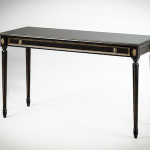 May include: A black rectangular console table with gold accents. The table has a single drawer with a gold knob and decorative gold detailing on the corners. The legs are slender and tapered, with a dark finish.