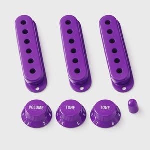 May include: A collection of purple guitar components, featuring three pickups, three control knobs marked "VOLUME" and "TONE", and a small purple switch tip. The knobs have white number markings. The parts are set against a light grey backdrop.