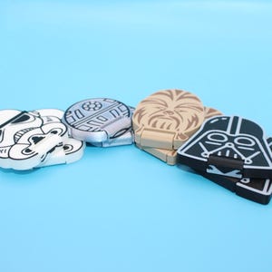 Pode incluir: Uma coleção de ímanes com o tema Star Wars. Os ímanes apresentam desenhos de um Stormtrooper, uma Estrela da Morte, Chewbacca e Darth Vader. Os ímanes são nas cores preto, branco e bege, sobre um fundo azul claro.
