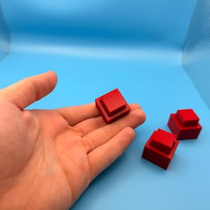 Peut inclure: Trois petits blocs carrés rouges à gradins. Un bloc est tenu dans une main, tandis que deux autres reposent sur une surface bleu clair. Les blocs ont un design géométrique simple et une finition mate.
