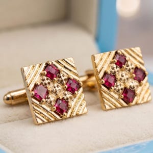 May include: A pair of square gold cufflinks featuring a textured diagonal striped pattern and four square-cut deep red gemstones set in a diamond formation on each piece. The cufflinks have a polished gold finish and a standard swivel toggle backing.
