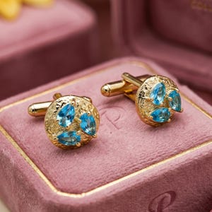 Peut inclure: Une paire de boutons de manchette dorés avec une surface circulaire texturée. Chaque bouton de manchette est orné de trois pierres précieuses bleu vif en forme de poire disposées en grappe. Le métal présente une finition martelée et irrégulière.