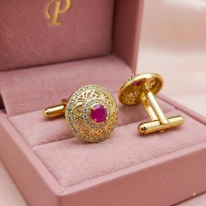 May include: A pair of gold-toned cufflinks featuring a circular design with an intricate filigree pattern, a border of small clear stones, and a central round red stone. The cufflinks have a polished metal finish and a standard toggle closure.