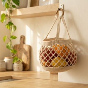 May include: A beige crochet hanging basket with a diamond-patterned mesh body and a solid circular base. The basket features two sturdy straps attached to a wooden ring, which is suspended from a wall hook, containing several fresh fruits.