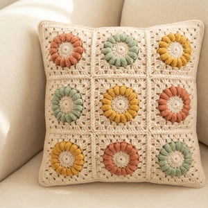 May include: A square crochet pillow cover featuring a cream-colored base with nine individual granny squares. Each square contains a raised, textured floral motif in muted shades of terracotta, sage green, and mustard yellow yarn.