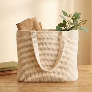 May include: A beige crochet tote bag with a rectangular shape and two handles. The bag is filled with brown paper and green leafy sprigs. The bag is sitting on a wooden table next to two books.