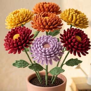 May include: A potted arrangement of crocheted flowers featuring yellow, orange, deep red, and light purple chrysanthemum-style blooms. Each flower has a textured center and is attached to a green stem with green leaves, all set in a terracotta pot.