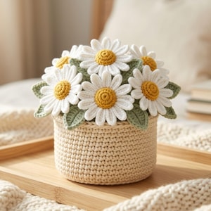 May include: A crochet daisy flower arrangement in a beige yarn basket. The flowers feature white petals and textured yellow centers, surrounded by sage green crocheted leaves. The basket is crafted with a dense, uniform stitch pattern in light beige.