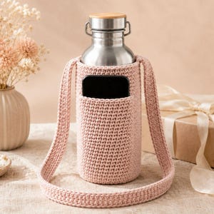 May include: A stainless steel water bottle with a wooden cap sits inside a light pink crocheted carrier with a shoulder strap. The carrier has a pocket for a phone. The background includes a vase with dried flowers and a wrapped gift.