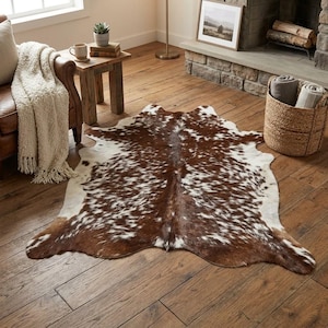 May include: A cowhide rug featuring a natural pattern of brown and white patches. The rug has an irregular, organic shape with a smooth texture, displaying a mix of dark brown, light brown, and white fur across its entire surface area.