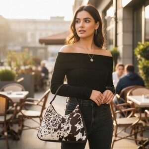 May include: A shoulder bag featuring a brown and white cowhide pattern with a dark brown braided handle. The bag has a curved top silhouette and dark brown trim along the edges, providing a structured look for carrying personal items.