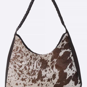 May include: A hobo-style shoulder bag featuring a brown and white cowhide pattern on the front and back panels. The bag is framed with dark brown leather trim and includes a single braided dark brown leather handle attached to the top sides.