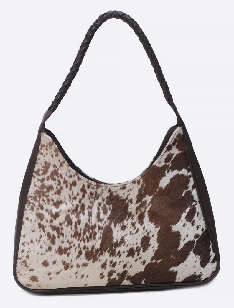 May include: A hobo-style shoulder bag featuring a brown and white cowhide pattern on the front and back panels. The bag is framed with dark brown leather trim and includes a single braided dark brown leather handle attached to the top sides.