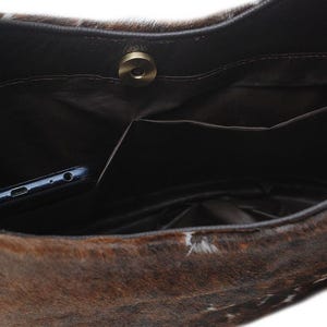 May include: A brown and white cowhide handbag interior featuring a dark brown fabric lining, a single internal slip pocket, and a circular magnetic snap closure. A dark blue smartphone is tucked inside the pocket of the bag.