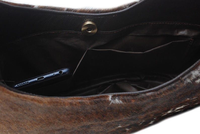 May include: A brown and white cowhide handbag interior featuring a dark brown fabric lining, a single internal slip pocket, and a circular magnetic snap closure. A dark blue smartphone is tucked inside the pocket of the bag.