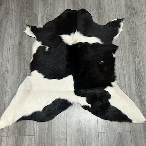 May include: A natural cowhide rug featuring a distinct black and white spotted pattern. The hide has an irregular, organic shape with a smooth, short-haired texture, showcasing large patches of solid black contrasted against bright white areas.