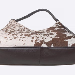 May include: A hobo-style handbag featuring a brown and white cowhide pattern on the upper section and a solid dark brown textured base. The bag includes a single braided dark brown handle attached to the top, providing a distinct contrast.