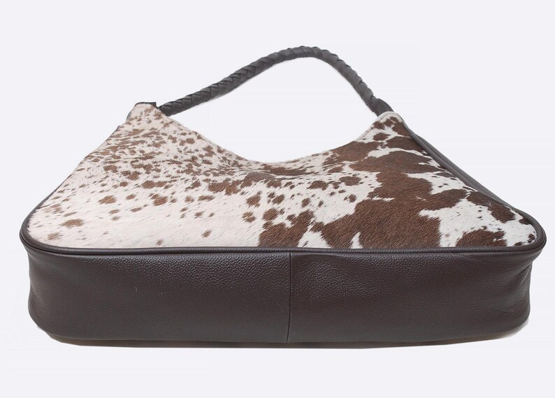 May include: A hobo-style handbag featuring a brown and white cowhide pattern on the upper section and a solid dark brown textured base. The bag includes a single braided dark brown handle attached to the top, providing a distinct contrast.