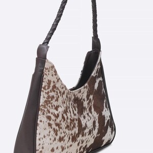 May include: A hobo-style shoulder bag featuring a brown and white cowhide print on the front and back panels. The bag includes solid dark brown leather side panels, a base, and a single braided leather handle attached to the top corners.