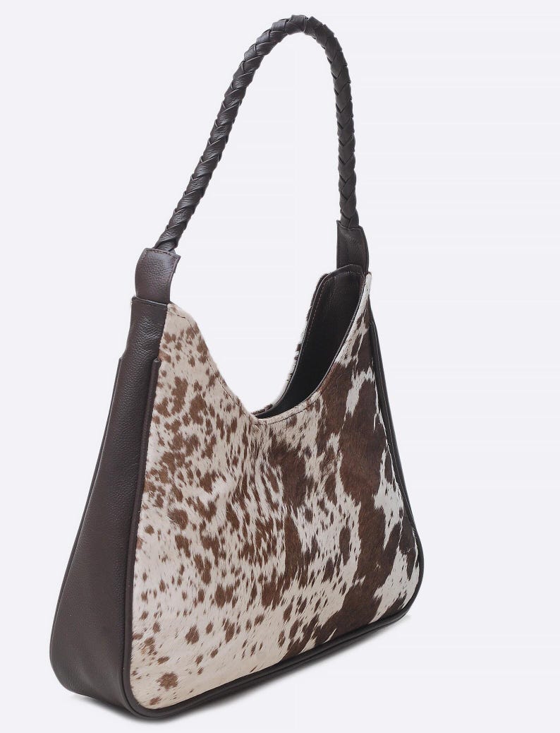 May include: A hobo-style shoulder bag featuring a brown and white cowhide print on the front and back panels. The bag includes solid dark brown leather side panels, a base, and a single braided leather handle attached to the top corners.