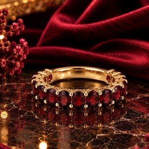 Könnte beinhalten: Ein goldener Eternity-Ring mit einer durchgehenden Reihe oval geschliffener, tiefroter Granat-Edelsteine, die in polierten Goldfassungen sitzen. Der Ring zeigt eine leuchtende, satte rote Farbe und ein metallisches Gold-Finish.