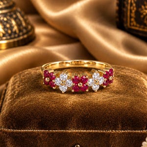 May include: A gold ring featuring a series of alternating flower-shaped clusters, each composed of either four round red gemstones or four round clear white gemstones, set in a continuous band design that wraps across the top of the ring.
