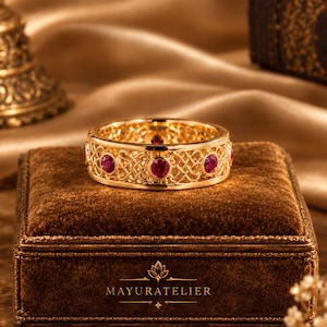 Könnte beinhalten: Ein goldener Bandring mit einem komplizierten, durchbrochenen Gitterdesign und mehreren runden, roten Edelsteinen, die in regelmäßigen Abständen eingefasst sind. Der Ring liegt auf einer braunen Samtschachtel mit dem goldenen Schriftzug MAYURATELIER.