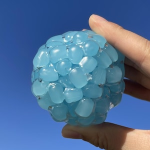 May include: A blue stress ball with a clear, gel-like exterior filled with small, light blue, pebble-shaped beads. The ball is held against a bright blue sky.