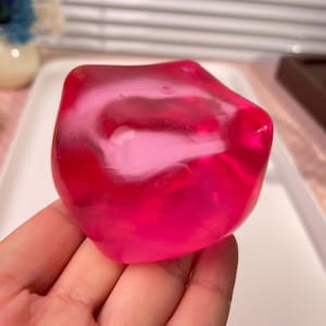May include: A translucent, vibrant hot pink object with a smooth, rounded, and irregular shape. The material appears glossy and semi-transparent, allowing light to pass through its solid, bulbous form, which fits comfortably within the palm of a hand.