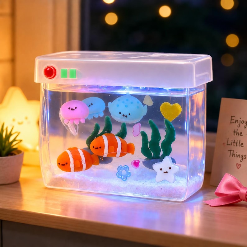 Może przedstawiać: Prostokątna lampka akwariowa z przezroczystego tworzywa sztucznego z wewnętrznym oświetleniem LED i miniaturowymi figurkami, w tym dwiema pomarańczowymi rybkami błazenkami, r&oacute;żową meduzą, niebieską płaszczką, białą gwiazdką i zielonymi algami.