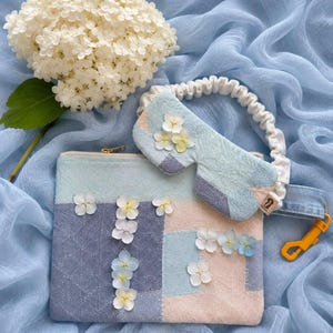 May include: A rectangular fabric pouch with a zipper closure and a matching sleep mask featuring an elasticized white headband. The items are decorated with a patchwork pattern in shades of light blue, dark blue, and beige, accented with small white flowers.
