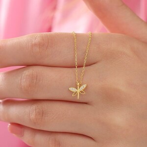 May include: A gold-coloured necklace with a dragonfly pendant. The dragonfly design includes small, sparkling details. The necklace is shown against a soft pink background, emphasising the jewellery's elegant design and delicate chain.