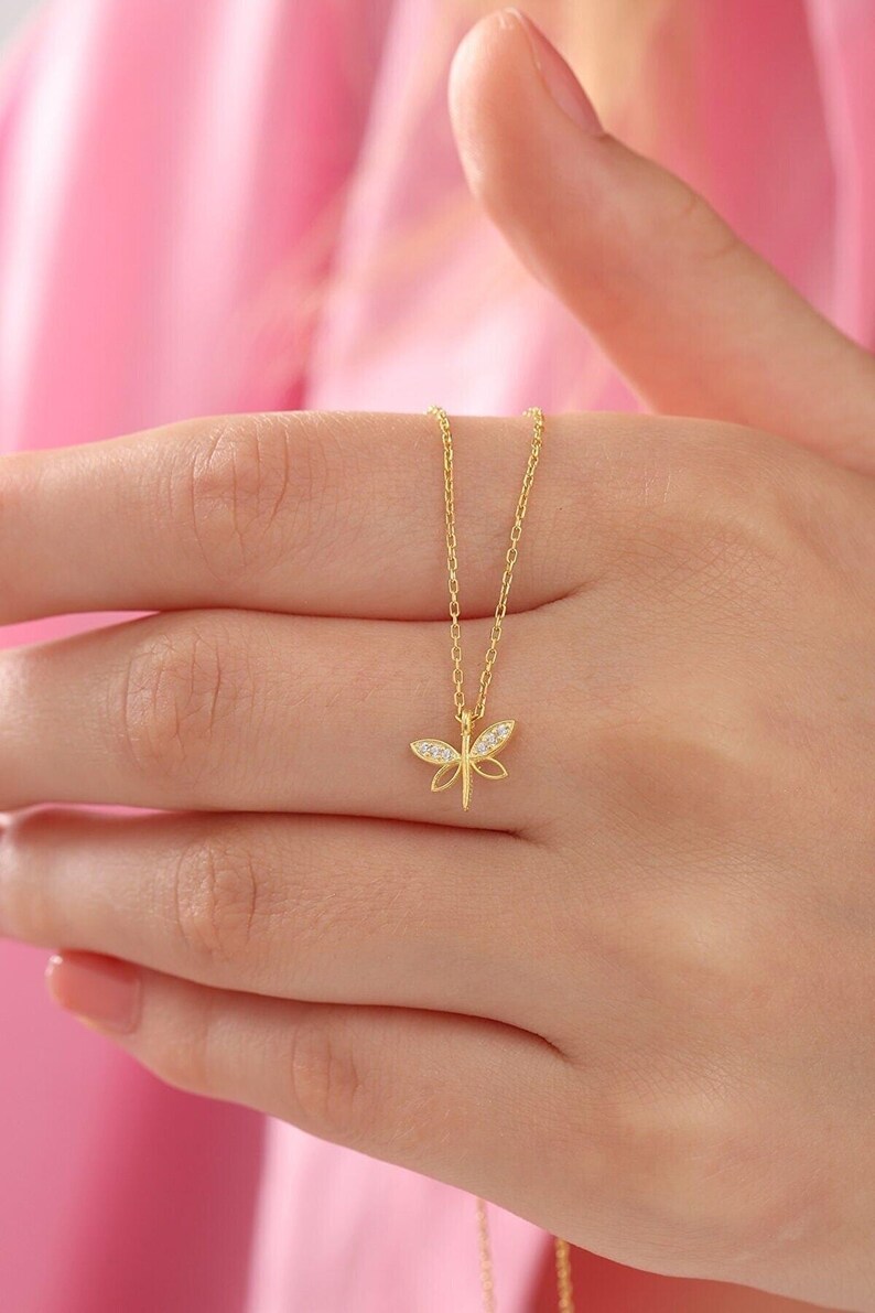 May include: A gold-coloured necklace with a dragonfly pendant. The dragonfly design includes small, sparkling details. The necklace is shown against a soft pink background, emphasising the jewellery's elegant design and delicate chain.