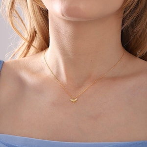 May include: A gold-coloured necklace with a dragonfly pendant. The dragonfly has small, sparkling details. The necklace is worn against a light skin tone and a blue top. The jewellery is a simple, elegant accessory.