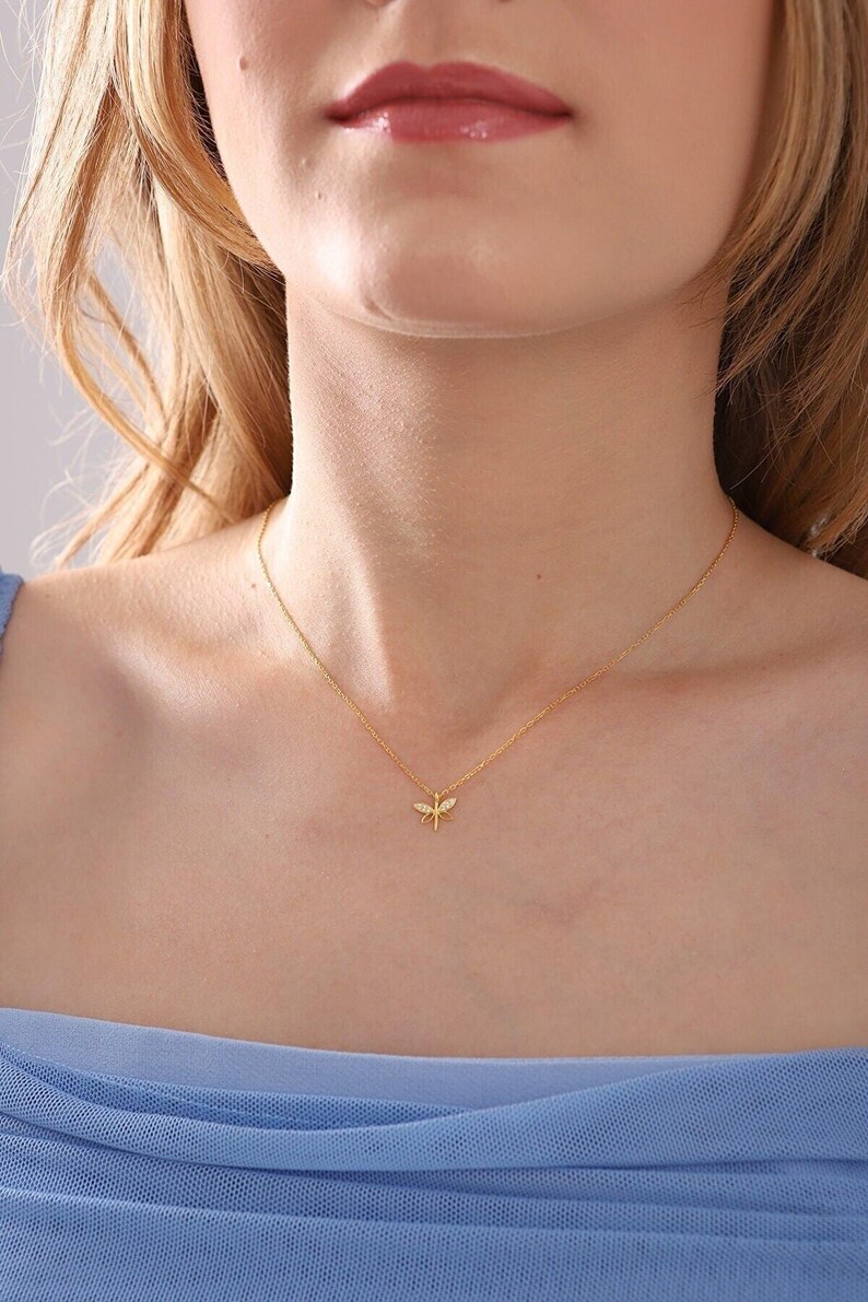 May include: A gold-coloured necklace with a dragonfly pendant. The dragonfly has small, sparkling details. The necklace is worn against a light skin tone and a blue top. The jewellery is a simple, elegant accessory.