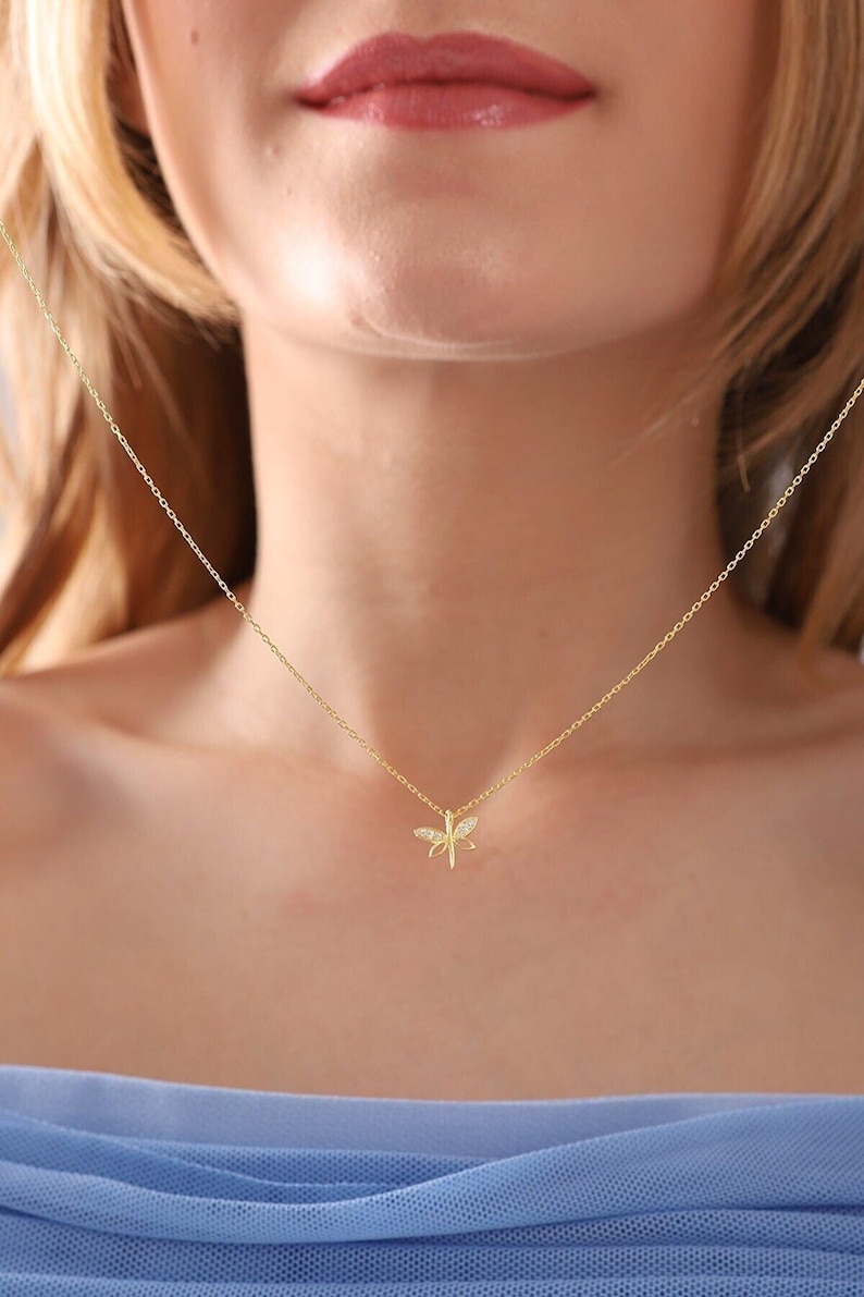 May include: A delicate gold necklace with a dragonfly pendant. The dragonfly has detailed wings and a small body, with what appears to be small gemstones. The necklace is worn around the neck.