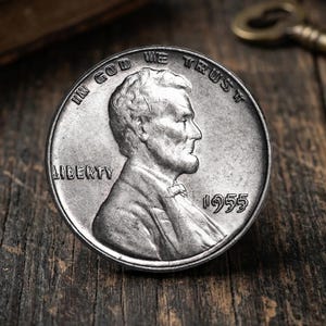 Puede incluir: Una moneda estadounidense de un centavo en tono plateado que presenta el perfil de Abraham Lincoln mirando hacia la derecha. La moneda muestra el texto IN GOD WE TRUST en el borde superior, LIBERTY a la izquierda y el año 1955 a la derecha.