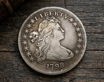 Może przedstawiać: Srebrna moneta z napisem "LIBERTY" i rokiem "1798". Moneta przedstawia profil kobiety otoczony gwiazdami. Leży na ciemnej drewnianej powierzchni, ukazując swój skomplikowany design i historyczne znaczenie.