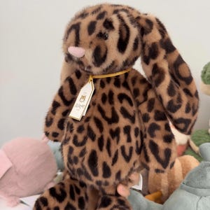 Op de afbeelding: Een pluche konijn met een luipaardprint. Het konijn heeft lange, slappe oren, een roze neus en een label om de nek. De vacht is zacht en de kleuren zijn bruin en zwart. Het speelgoed is ontworpen voor kinderen.
