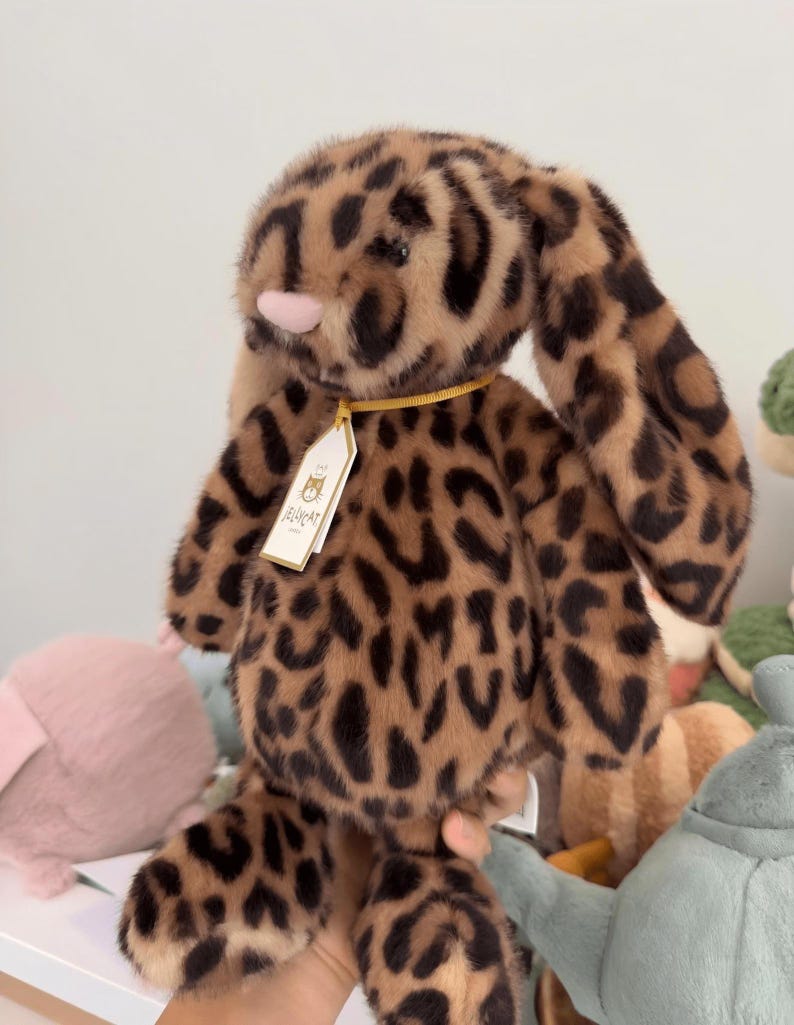 Op de afbeelding: Een pluche konijn met een luipaardprint. Het konijn heeft lange, slappe oren, een roze neus en een label om de nek. De vacht is zacht en de kleuren zijn bruin en zwart. Het speelgoed is ontworpen voor kinderen.