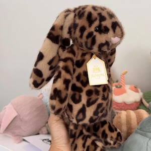 Op de afbeelding: Een pluche konijn met een luipaardprint in bruin- en beigetinten. Het konijn heeft lange, slappe oren en een roze neus. Een label is aan het oor bevestigd. Andere knuffels zijn op de achtergrond te zien.