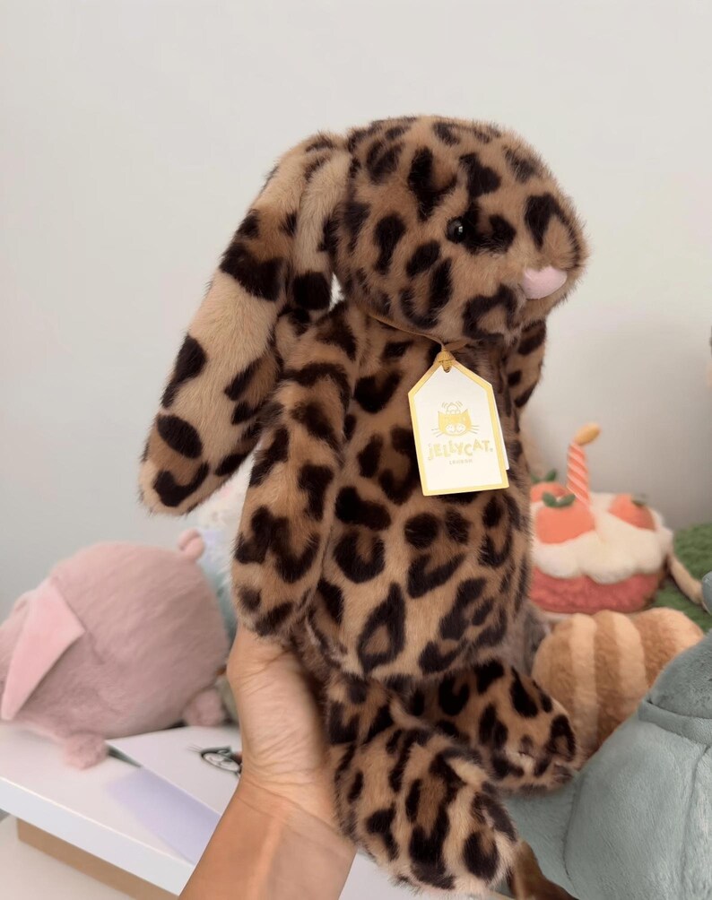 Op de afbeelding: Een pluche konijn met een luipaardprint in bruin- en beigetinten. Het konijn heeft lange, slappe oren en een roze neus. Een label is aan het oor bevestigd. Andere knuffels zijn op de achtergrond te zien.