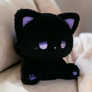 Könnte beinhalten: Ein schwarzes Plüschkätzchen mit lila Akzenten an Ohren und Pfoten. Die Katze hat gestickte Augen und eine kleine Nase. Das Kätzchen sitzt auf einer weichen, hellfarbigen Oberfläche.