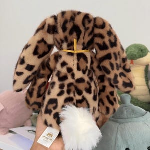 Op de afbeelding: Een pluche konijn met een luipaardprint in bruin- en zwarttinten. Het konijn heeft lange, slappe oren, een witte, pluizige staart en een gele strik. Het label luidt "Jellycat London".