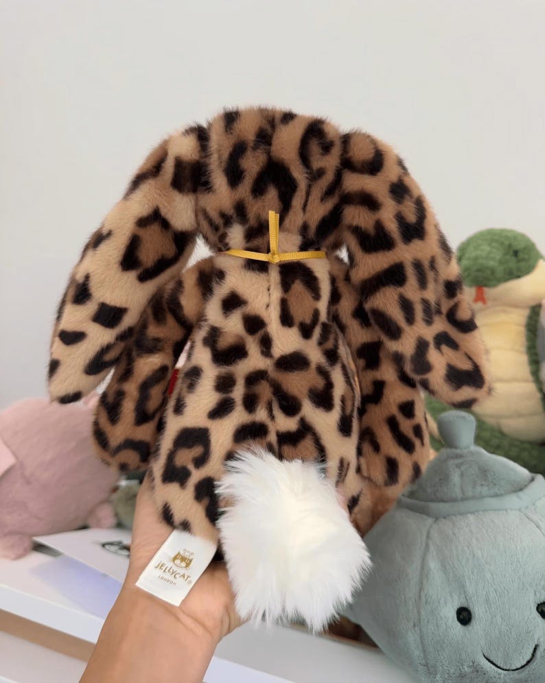 Op de afbeelding: Een pluche konijn met een luipaardprint in bruin- en zwarttinten. Het konijn heeft lange, slappe oren, een witte, pluizige staart en een gele strik. Het label luidt "Jellycat London".