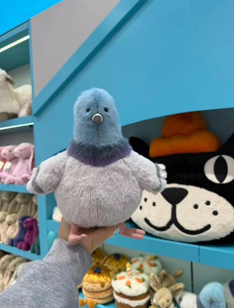 K&ouml;nnte beinhalten: Ein Pl&uuml;schpigeon mit blauem Kopf und grauem K&ouml;rper. Das Spielzeug hat einen lila Kragen und kleine rosa F&uuml;&szlig;e. Im Hintergrund sind weitere Pl&uuml;schtiere auf Regalen zu sehen, darunter eine schwarze Katze mit orangefarbenem Hut.